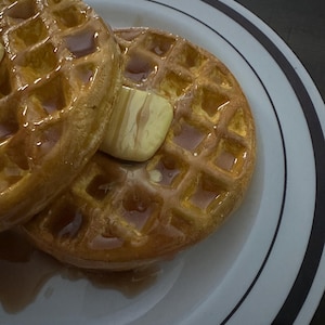 May include: Two golden-brown waffles, topped with a pat of butter and drizzled with syrup, sit on a white plate with black and gray stripes. The waffles have a square grid pattern and are glistening with syrup.