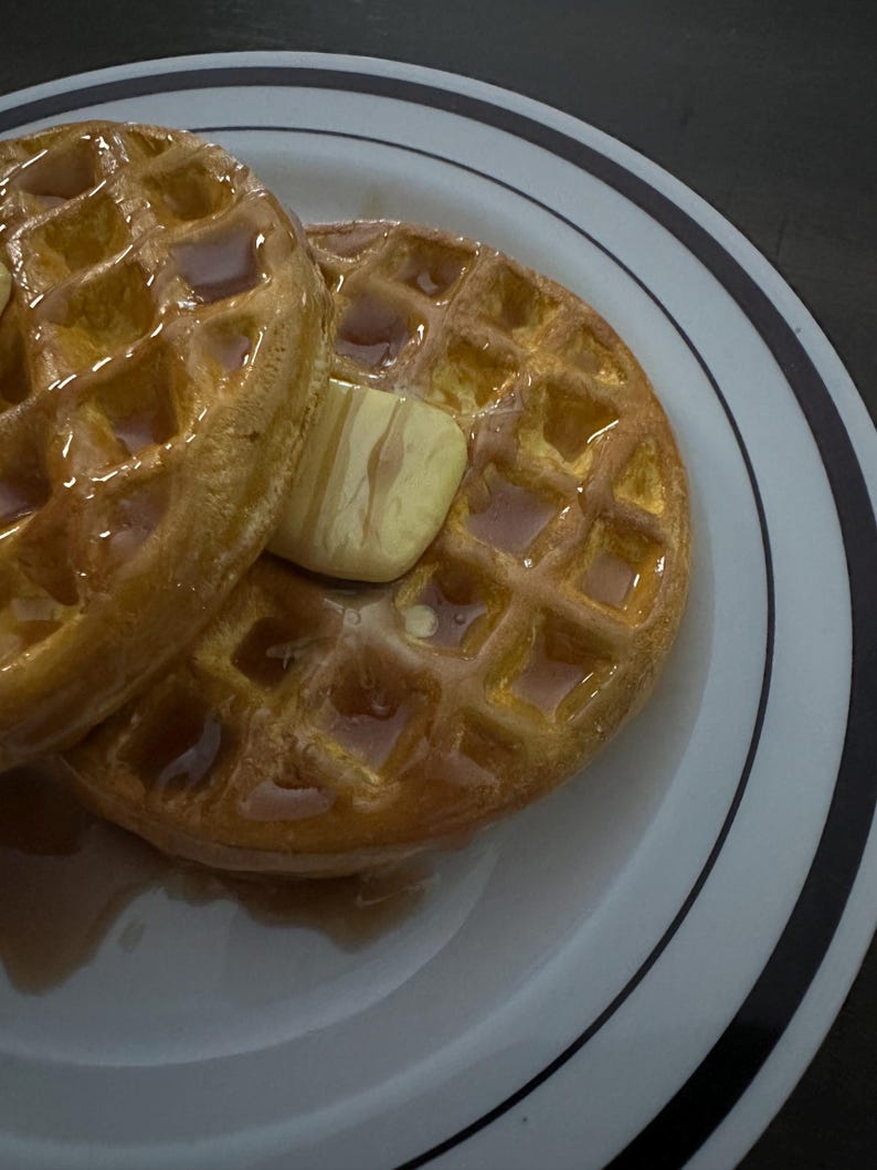 May include: Two golden-brown waffles, topped with a pat of butter and drizzled with syrup, sit on a white plate with black and gray stripes. The waffles have a square grid pattern and are glistening with syrup.
