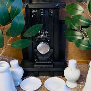 Op de afbeelding: Een miniatuur zwarte open haard met een ronde spiegel en decoratieve elementen. Verschillende witte keramische stukken, waaronder kleine borden en diverse gevormde containers, zijn voor de open haard geplaatst. Groene bladeren voegen een natuurlijk tintje toe.