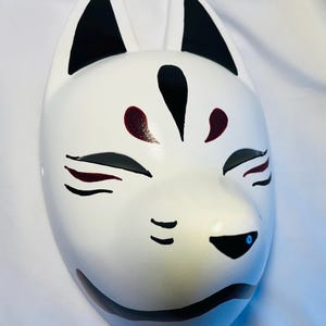 以下が含まれることがあります： 黒色の耳、目、鼻が付いた白い狐のマスク。マスクは目の周りに赤い模様があり、顔の中央に黒い線が引かれている。