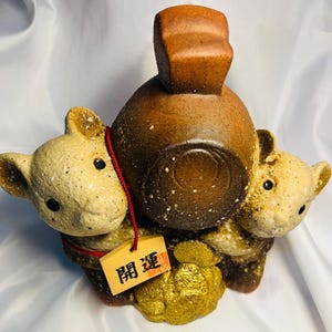 以下が含まれることがあります： 2匹のクマのような姿が、四角い上部を持つ茶色の容器を挟んだセラミック彫刻。クマはベージュ色で、黒い目と赤い紐が付いています。日本語の文字が書かれた小さな木の札が紐からぶら下がっています。台座は金色です。