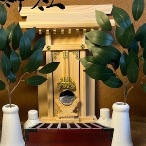 May include: A small wooden shrine with a red wooden box in front of it. The shrine has a gold trim and a mirror inside. The box has the Japanese characters "奉納" written on it. There are two white ceramic vases with green leafy plants on either side of the shrine.