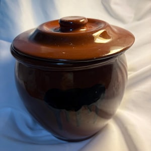 Puede incluir: Un tarro de cerámica marrón de forma redonda con tapa a juego. El tarro presenta un acabado brillante y un patrón de esmalte de goteo de color negro oscuro que recorre el lateral del recipiente. La tapa tiene un pequeño pomo central.