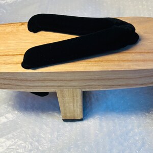 May include: A pair of wooden geta sandals with a black velvet strap. The sandals have a raised wooden platform and a black rubber sole.