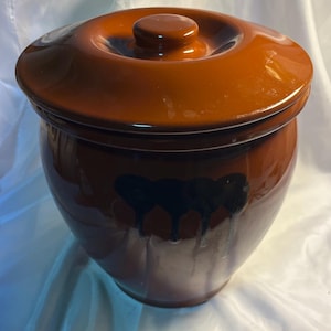 May include: A brown ceramic crock with a matching lid. The crock has a glossy finish and a dark blue decorative design. The lid has a small knob handle. The crock is likely used for food storage.