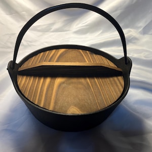 Könnte beinhalten: Ein schwarzer gusseiserner Topf mit einem Holzdeckel und einem gebogenen Griff. Der Deckel besteht aus hellbraunem Holz mit einem natürlichen Maserungsmuster. Der Topf ist zum Kochen bestimmt.