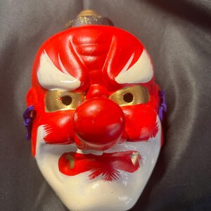 May include: A red and white Japanese Noh mask with gold accents. The mask features a prominent red nose, white eyebrows, and gold-lined eye holes. Purple cords are attached to the sides. The mask is a traditional Japanese art form.