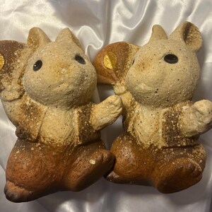 以下が含まれることがあります： 大きな耳と翼を持つ、様式化された動物の2つの陶器製置物。フィギュアは主にベージュ色で、茶色のアクセントと翼に金の円があります。黒い目を持ち、インタラクションしているかのようにポーズをとっています。