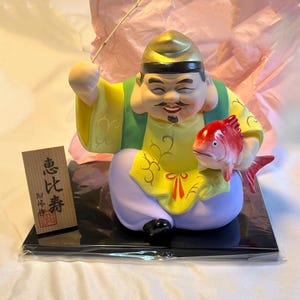 May include: A ceramic figurine of a man holding a red fish. He is wearing a yellow and green robe with a gold hat. The figurine is sitting on a black base. The text on the label reads "恵比寿", which is the Japanese name for the god of prosperity and good fortune.