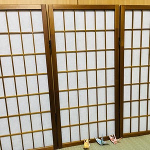 May include: A three-panel folding room divider featuring a brown wooden frame with a grid pattern and white translucent paper panels. Three small, colourful origami paper cranes are positioned in front of the base of the centre panel.