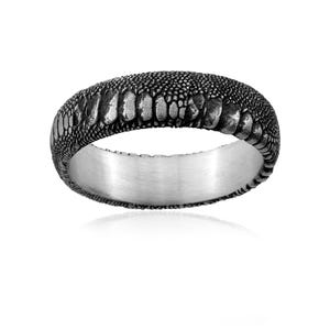 Peut inclure: Une bague en argent avec un anneau texturé gris foncé. La surface de l'anneau présente un motif rappelant la peau de reptile. La bague est posée sur un fond blanc, soulignant son design unique et son éclat métallique.