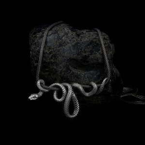 Può includere: Una collana di serpente d'argento con un design testurizzato e avvolto. La collana è drappeggiata su una roccia grigio scuro.
