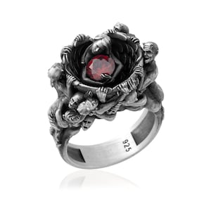 Pode incluir: Anel de prata com uma pedra vermelha central, rodeada por figuras esculpidas. O anel tem um acabamento detalhado e antigo, com a marca "925" visível na banda. O design é intrincado e artístico.
