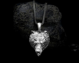 May include: A silver bear head pendant necklace on a black chain. The pendant is detailed and realistic, with fur and a sharp snout. The necklace is resting on a dark gray rock.