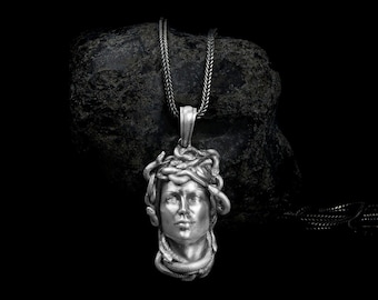 May include: A silver pendant necklace featuring a detailed sculpture of Medusa's head with snakes for hair. The pendant is hanging from a silver chain and resting on a dark gray rock.