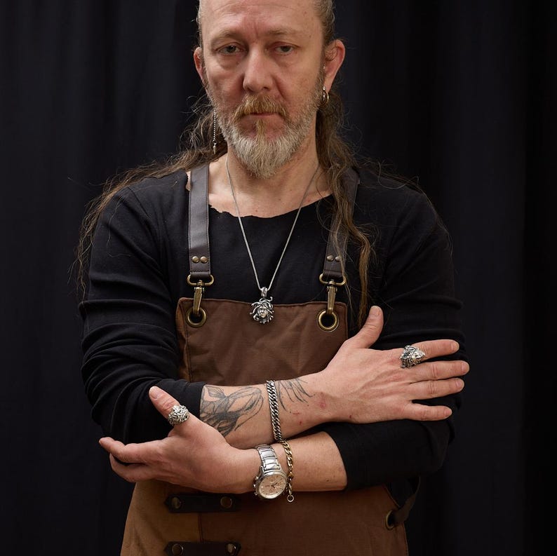 May include: A man with long, dark hair and a beard is wearing a brown leather apron with a silver pendant necklace. He is wearing a silver ring on his right hand and a silver bracelet on his left wrist. He is also wearing a silver watch on his left wrist.