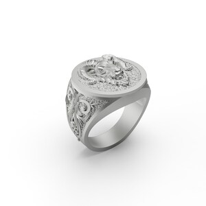May include: A silver ring with an ornate design featuring a ram's head on the top. The ring has a wide band with intricate details.