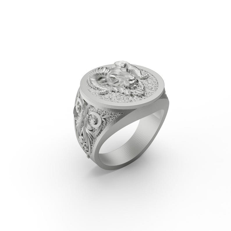 May include: A silver ring with an ornate design featuring a ram's head on the top. The ring has a wide band with intricate details.