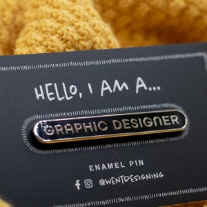 May include: A black business card with the text "HELLO, I AM A... GRAPHIC DESIGNER" and an enamel pin. The pin is silver with the words "GRAPHIC DESIGNER" in black. The card is on a yellow knitted fabric.