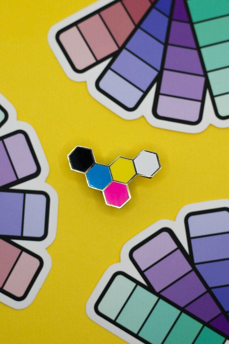 May include: A colorful enamel pin in a hexagonal design, featuring black, blue, pink, yellow, and white segments, set against a bright yellow background. Surrounding the pin are several color palette stickers with various shades of purple, pink, and green.