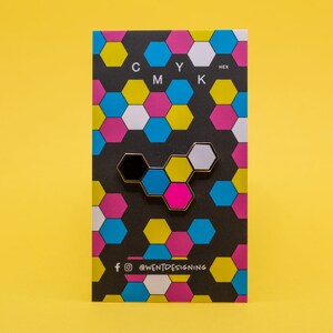 May include: A colorful enamel pin in a honeycomb design, featuring black, white, pink, blue, and yellow hexagons. The pin is on a black card with a repeating pattern of the same colors and the letters "CMYK" at the top.
