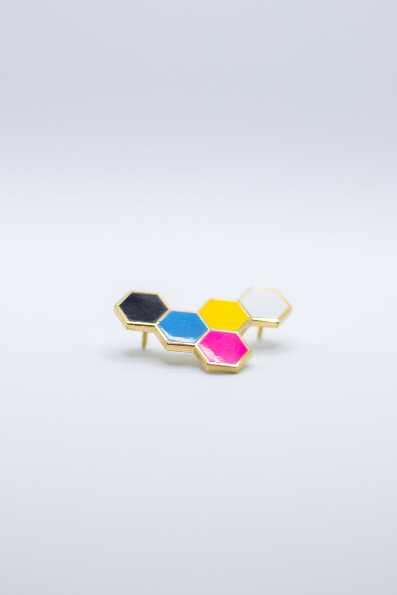 May include: A gold-toned pin with a honeycomb design. Each hexagon is a different color: black, blue, yellow, pink, and white. The pin has two posts on the back for secure attachment.