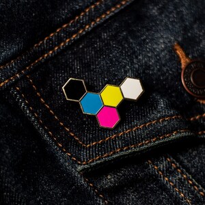May include: A colorful enamel pin in a geometric design, featuring six connected hexagons. The pin has a gold-colored border and is filled with black, blue, yellow, pink, and white enamel. It is set against a dark blue denim jacket.