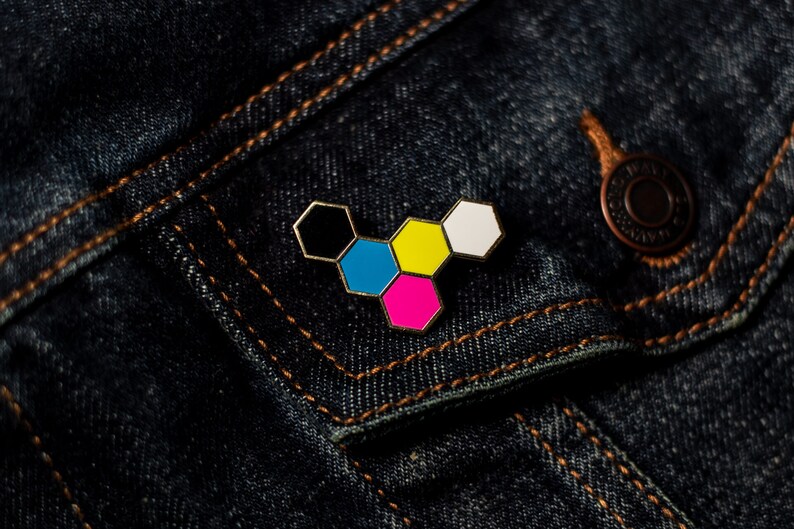 May include: A colorful enamel pin in a geometric design, featuring six connected hexagons. The pin has a gold-colored border and is filled with black, blue, yellow, pink, and white enamel. It is set against a dark blue denim jacket.