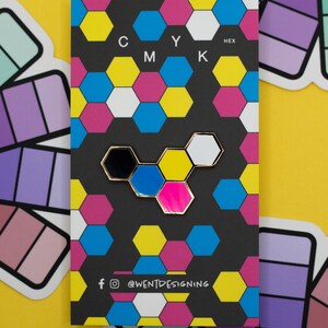 May include: A black card with a colorful hexagonal pattern and the letters CMYK and HEX. A gold-rimmed pin with black, blue, pink, and white hexagons is attached. The card has social media icons and the text "@WENTDESIGNING".