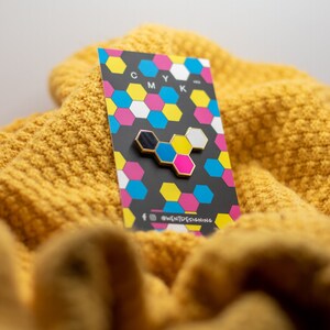 May include: A gold-edged enamel pin featuring a CMYK design, with black, blue, yellow, pink, and white hexagonal shapes. The pin is displayed on a card with a matching CMYK pattern, resting on a textured yellow knit fabric.