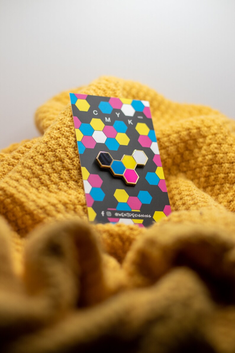 May include: A gold-edged enamel pin featuring a CMYK design, with black, blue, yellow, pink, and white hexagonal shapes. The pin is displayed on a card with a matching CMYK pattern, resting on a textured yellow knit fabric.
