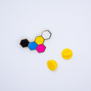 May include: A colorful enamel pin featuring a geometric design of connected hexagons in black, blue, pink, yellow, and white, with a gold border. Two yellow pin backs are also visible on a white background.