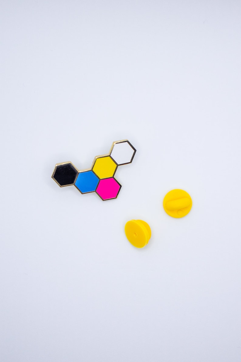 May include: A colorful enamel pin featuring a geometric design of connected hexagons in black, blue, pink, yellow, and white, with a gold border. Two yellow pin backs are also visible on a white background.