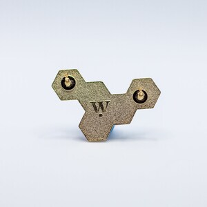 May include: A gold-colored, geometric pin with a honeycomb design. The pin features three hexagonal shapes connected together, with a small "W" logo in the center. Two pin backs are visible. The pin is set against a white background.