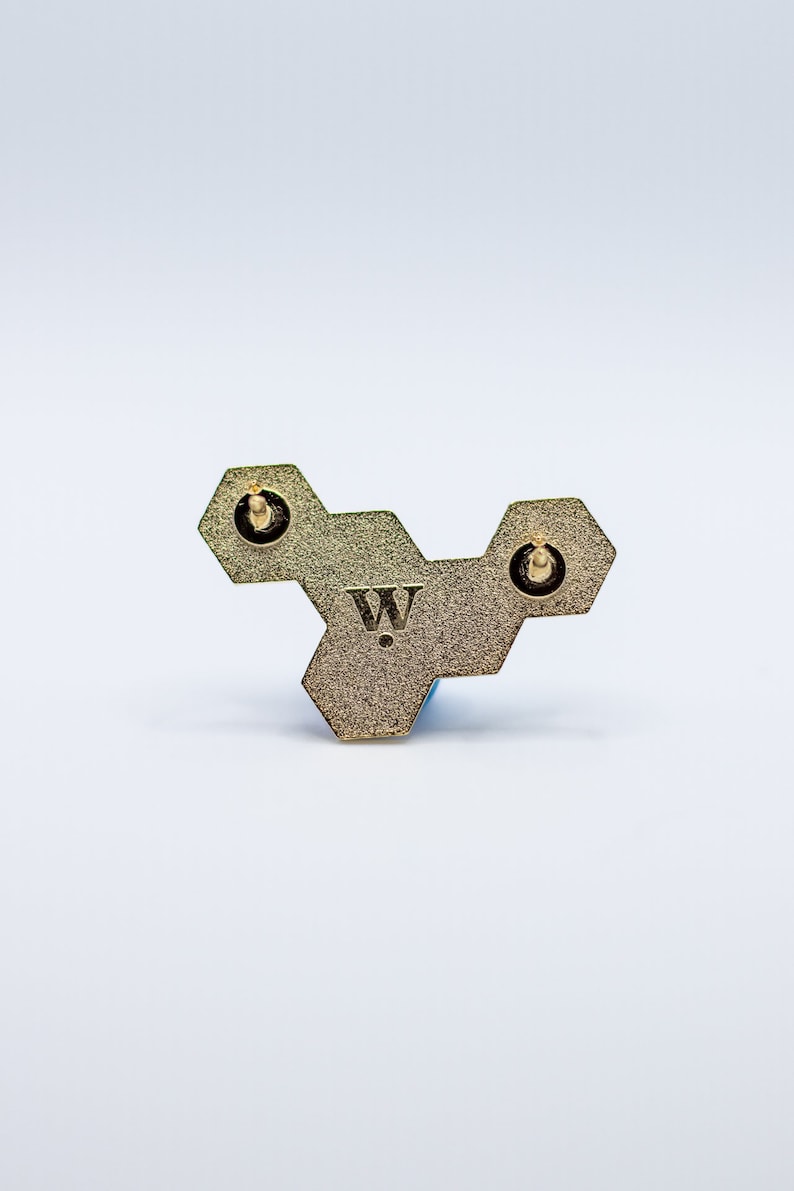 May include: A gold-colored, geometric pin with a honeycomb design. The pin features three hexagonal shapes connected together, with a small "W" logo in the center. Two pin backs are visible. The pin is set against a white background.