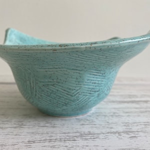 May include: A light blue ceramic bowl with a textured, patterned surface. The bowl has a wide, shallow shape with a slightly flared rim.