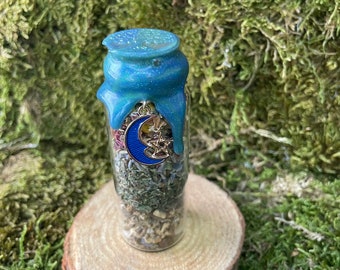 May include: A small glass bottle filled with dried herbs and flowers, sealed with a blue and green glitter resin top. A gold charm with a blue crescent moon and a star hangs from the bottle.