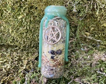May include: A small glass bottle with a green, glittery, dripping top. The bottle is filled with dried herbs and a silver pentagram charm. The bottle is sitting on a bed of green moss.