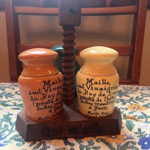 Puede incluir: Un juego de vinagre y aceite vintage con dos tarros de cerámica y una prensa de tornillo de madera. Los tarros están etiquetados con texto francés y tienen tapas naranjas y amarillas. El juego está sobre una base de madera.