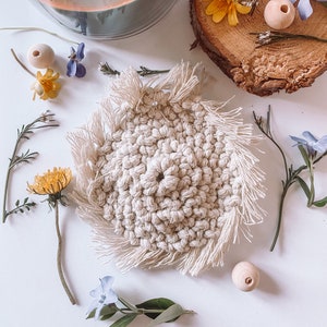 Könnte beinhalten: Ein beigefarbener Makramee-Untersetzer mit Fransen ist von getrockneten Blumen, Holzperlen und grünen Zweigen umgeben. Eine brennende Kerze in einem Glas und eine Holzscheibe tragen zur natürlichen Ästhetik bei. Der Untersetzer ist handgefertigt.