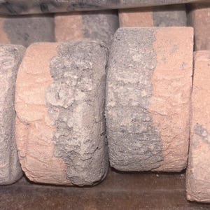 May include: A collection of cylindrical, weathered stones. Each stone is a mix of pink and gray, with a rough, textured surface. The stones appear to be stacked or arranged together, possibly for landscaping or construction purposes.