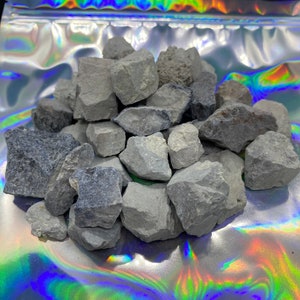 May include: A pile of gray, irregularly shaped rocks. The rocks vary in size and texture, with some appearing smoother than others. The rocks are in a silver, iridescent bag. The background is a shiny, holographic surface.