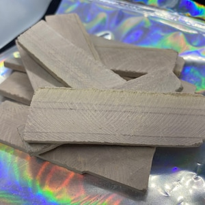 May include: A collection of light gray, rectangular, textured pieces, possibly wood or stone, are stacked and scattered. The pieces have a rough, natural appearance with visible grain or striations. The background includes a shiny, iridescent pouch.