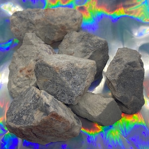 May include: A collection of rough, gray rocks with varying sizes and textures. The rocks have a natural, earthy appearance, with subtle color variations. The background is a shiny, iridescent surface, creating a contrast with the rocks' muted tones.
