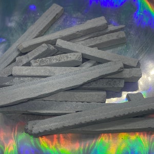 May include: A collection of grey slate chalk sticks, rectangular in shape, with rough, textured surfaces. The chalk pieces are scattered on a reflective, iridescent surface. The chalk sticks appear to be new and unused, ready for use in education or art.