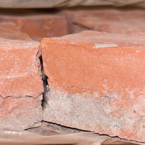May include: A collection of rectangular, red-orange bricks with a weathered appearance. The bricks have a rough, textured surface and a light-colored base. They are stacked together, partially visible within a clear plastic wrapping.
