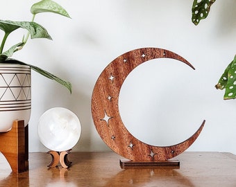 Op de afbeelding: Een houten halve maan met uitgesneden sterren, een witte bol op een houten standaard en een plant in een pot met groene bladeren in een witte en gouden pot op een houten standaard. Alle items staan op een houten tafel.