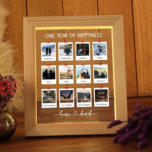 Può includere: Una cornice in legno con sfondo bianco e una griglia di 12 piccole foto. Le foto sono disposte in formato calendario, con ogni mese etichettato. Il testo "ONE YEAR OF HAPPINESS" è in cima alla cornice. I nomi "Laura & David" sono in fondo alla cornice.