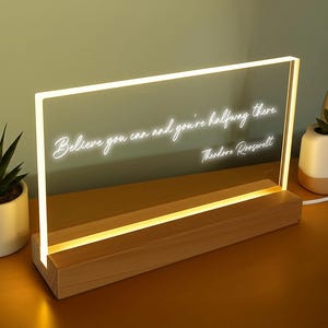 May include: A rectangular LED light with the quote "Believe you can and you're halfway there." The light has a wooden base and a clear acrylic panel. The quote is in a cursive font. The light is on a wooden surface.