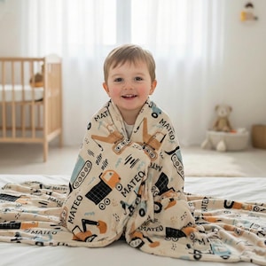 Può includere: Una coperta beige per bambini con un motivo di veicoli da costruzione e il nome "MATEO" stampato sopra. La coperta, con illustrazioni arancioni, blu e nere, è avvolta attorno a un bambino sorridente.